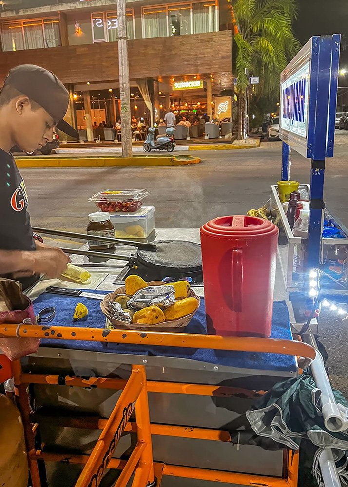 A vendor holds a freshly made Marquesita, a traditional Yucatecan dessert, in Playa Del Carmen, Mexico. The setting is a busy street with a food vendor stall in the background, and the weather is warm and sunny.