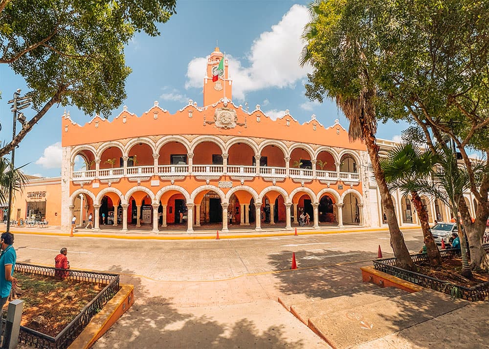 The Mérida Municipal Palace in Mérida, Mexico, showcasing its beautiful colonial architecture with arches and a clock tower. The weather is sunny with a few clouds, and people are seen walking around the square.