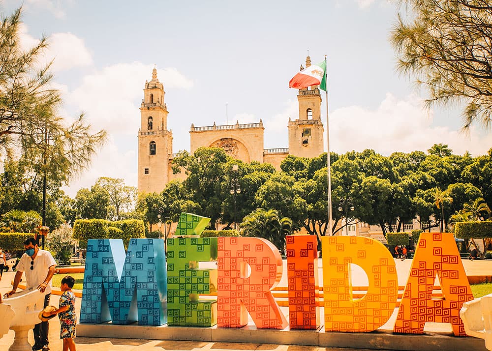 A colorful "MÉRIDA" sign in Mérida, Mexico, with the historic cathedral and lush greenery in the background. The sunny weather and clear sky highlight the vibrant culture and heritage of the city.