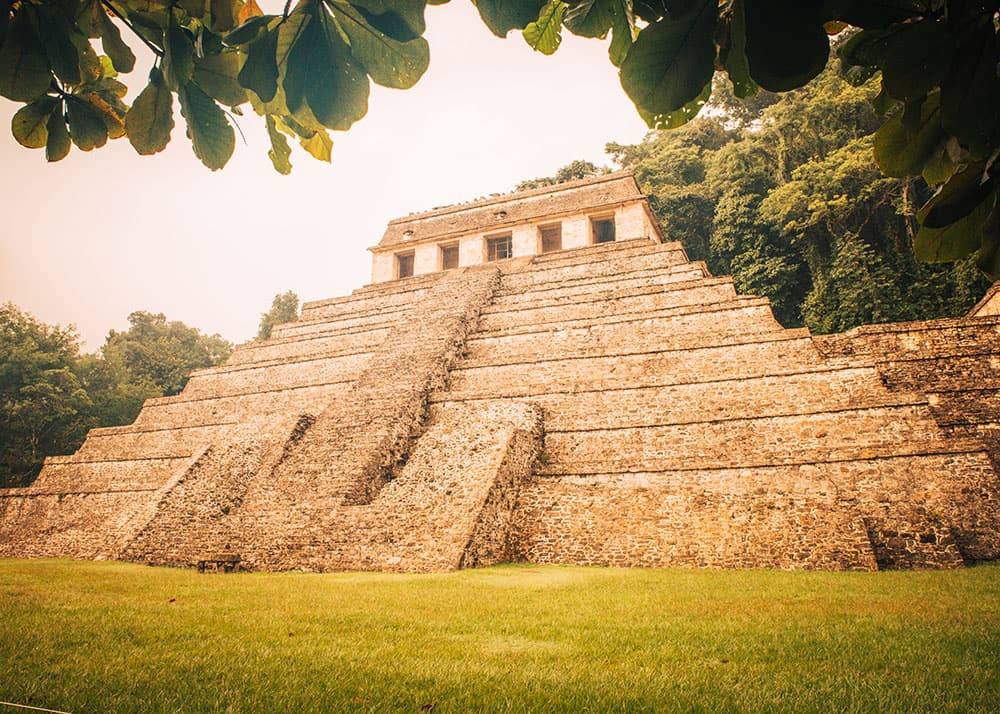 The grand pyramid at Palenque ruins in Chiapas, Mexico, rises against a backdrop of lush greenery and a cloudy sky. The well-preserved stonework and expansive lawn create a majestic and historical scene.