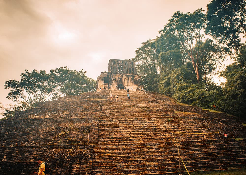 Tourists climb the steps of a large pyramid at the Palenque ruins in Chiapas, Mexico, with trees and an overcast sky in the background. The massive stone structure stands as a testament to the ancient civilization that built it.