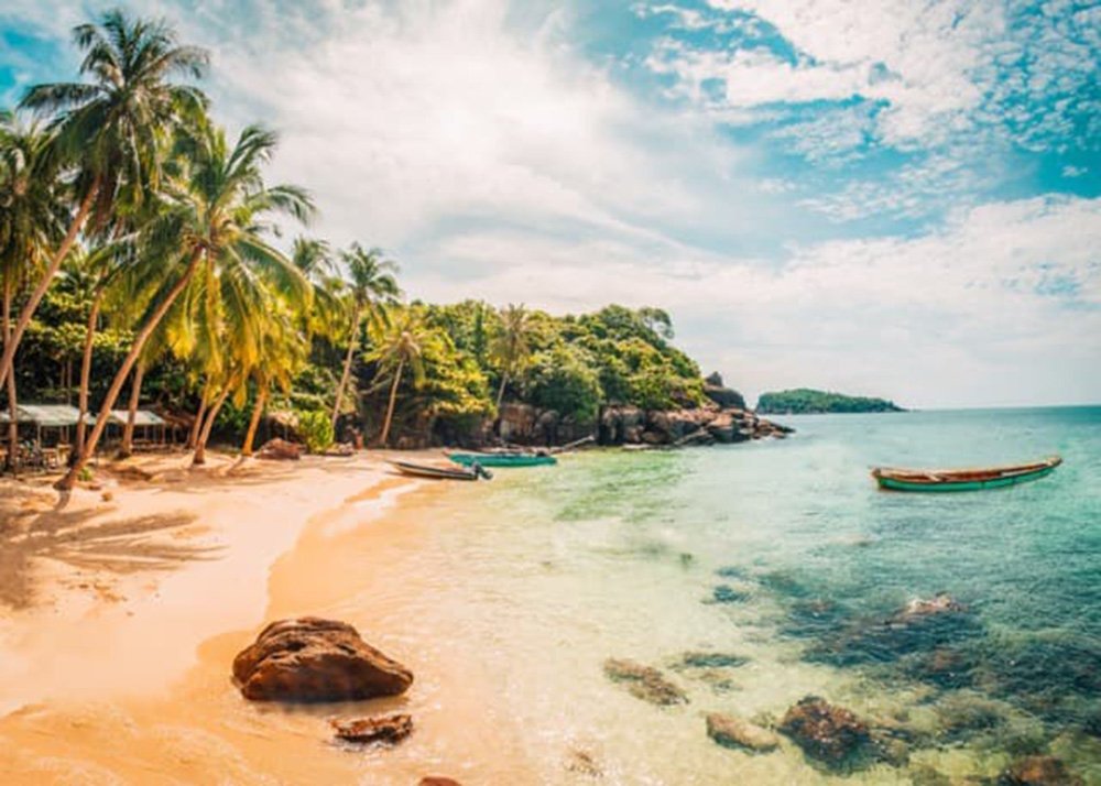 The azure blue sea reaches Phu Quoc. The beach is covered in white sand and small boulders. Palm trees fill the coastline.