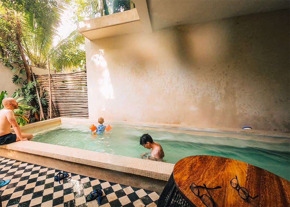 A family enjoys a private pool in Tulum, Mexico, with one child swimming and an adult relaxing by the poolside. The weather is warm, and the area is surrounded by tropical greenery, providing a secluded retreat.