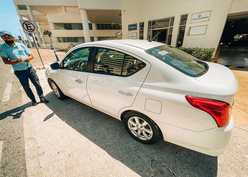 A family stands beside a rental car in Cancun, Mexico, with the car rental office in the background. The weather is clear and sunny, and the family appears excited and ready to embark on their road trip.