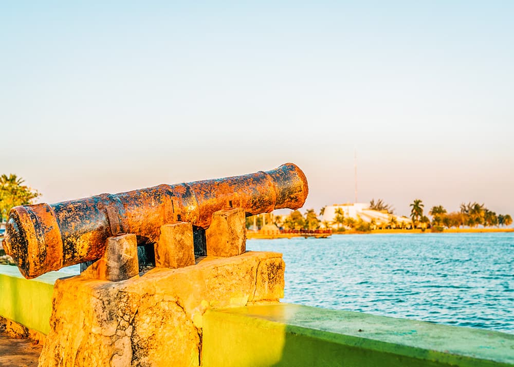 A rusted cannon overlooks the calm waters of the bay in Chetumal, Mexico, during a sunny day. The promenade and distant buildings are bathed in warm sunlight, adding to the historical ambiance.