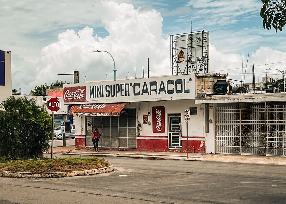 A small, local shop named "Mini Super Caracol" in Chetumal, Mexico, with a Coca-Cola sign and a person standing outside. The weather is partly cloudy, and the shop adds to the authentic local vibe.