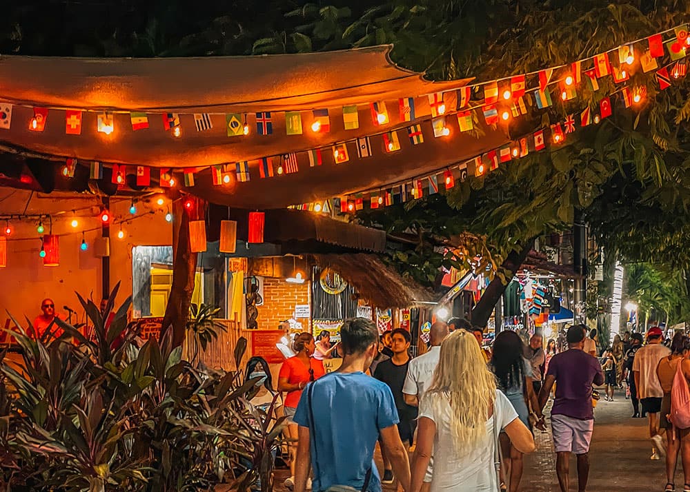 Another view of a street in Playa Del Carmen, Mexico, highlighting the relaxed atmosphere with people walking, biking, and exploring the area. It's evening and the street is filled with strings of lights that are lit up.