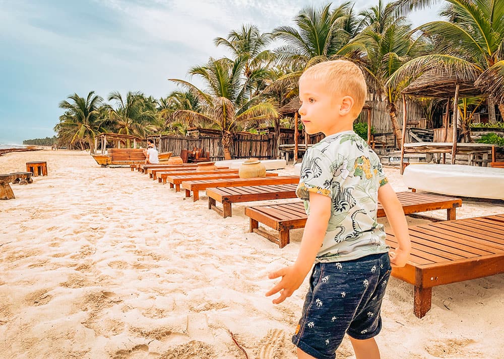 A young boy walks along the sandy beach at a beach club in Tulum, Mexico, with rows of wooden loungers and palm trees in the background. The weather is warm and inviting, perfect for a family day at the beach.