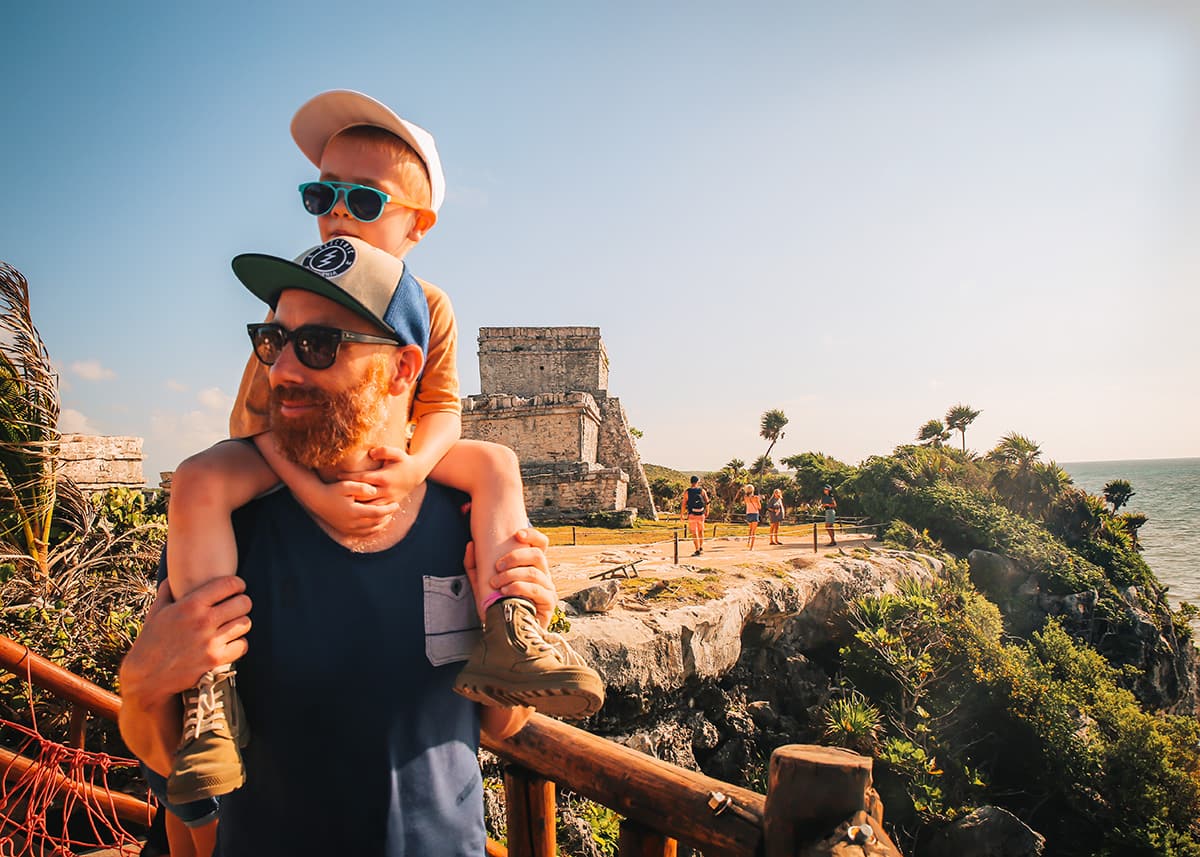 A boy with a white cap and blue sunglasses on the shoulders of his father in the water. His father wears a blue and white cap and brown sunglasses. They are exploring the ruins in Tulum.