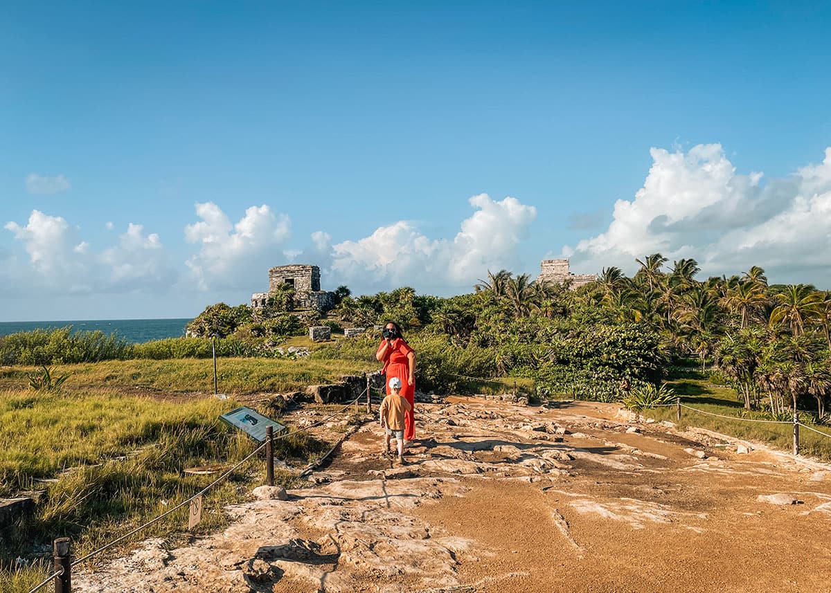 A woman in a red jumpsuit is gazing at the El Castillo (at the Tulum Ruins), while her toddler son runs up to her. It's a warm day with beautiful blue skies