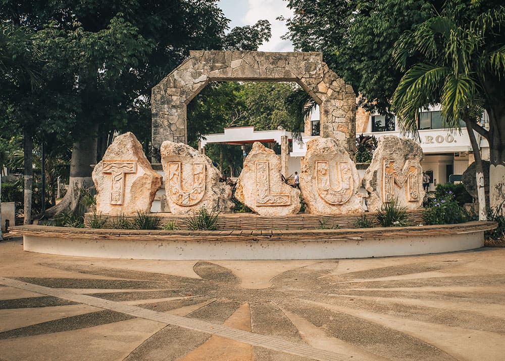 A large stone sign spelling out "Tulum" in Tulum, Mexico, surrounded by trees and local buildings. The weather is clear, and the sign serves as a popular photo spot for visitors.