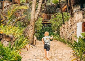 A young boy runs through the sandy beach at a beach club in Tulum, Mexico, with the ocean waves in the background. The weather is warm and sunny, perfect for beach activities.