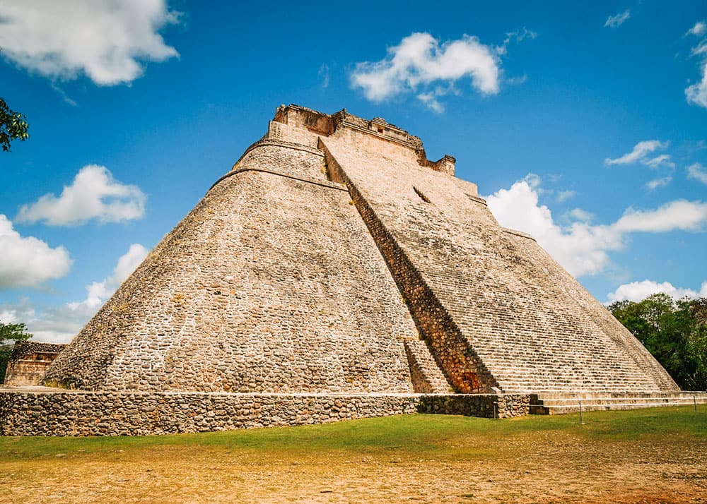The imposing Pyramid of the Magician at Uxmal, Mexico, stands tall against a blue sky with scattered clouds. The well-preserved stone structure showcases the architectural prowess of the ancient Maya civilization.