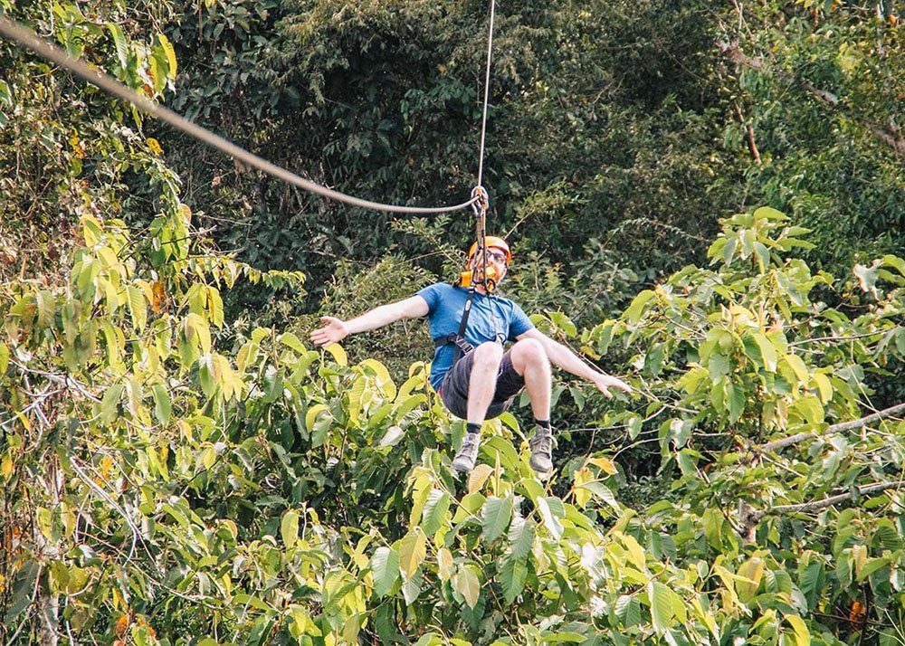 A man spreads his arms in excitement while ziplining through the dense jungle canopy. The weather is sunny, and the thrill of the adventure is captured in his joyful expression.