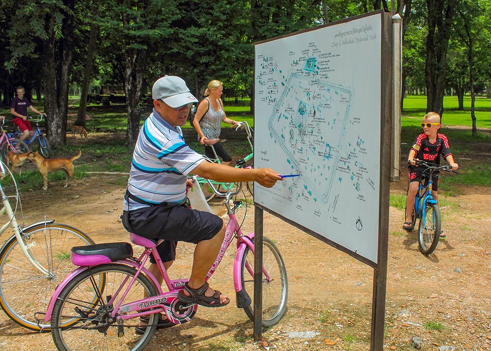 A tour guide on a pink bike points at a map of Sukhothai Historical Park with a blue pen, while his group looks on.