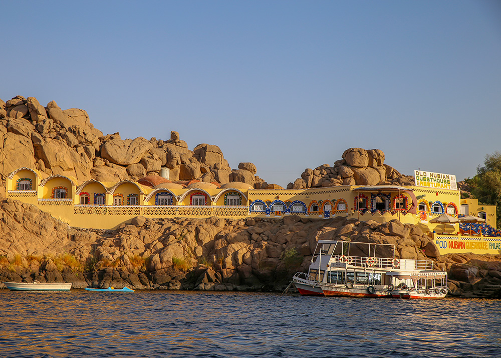 The Old Nubian Guest House & Restaurant as seen from the water. A motorboat is docked in front of it. The hotel looks as if it's part of the rocks that's surrounding it. The name of the place is painted in different colors. And nubian details are painted around every window.