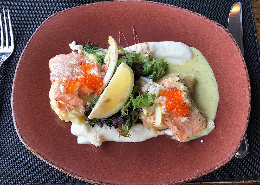 A beautiful pink/terracota colored plate in a restaurant is beautifully plated with a ligh green foamy sauce underneath two pieces of Arctic Char with salmon eggs on top as well. In the middle a few pieces of lettuce and 2 slices of lemon.