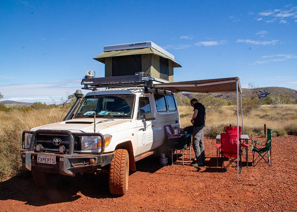 A man is cooking underneath the awning of a troopy campervan on a gravel campground