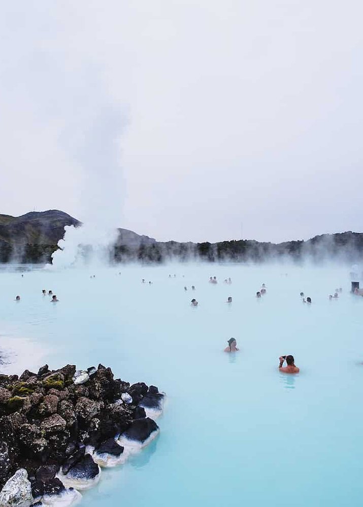 People are enjoying the Blue Lagoon. Lava fields can be seen surrounding the geothermal pool. Steam can be seen coming from the water.