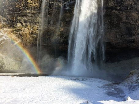 A waterfall in Iceland cascades into partially frozen water. The water is covered in snow and a rainbow can be seen right next to the waterfall