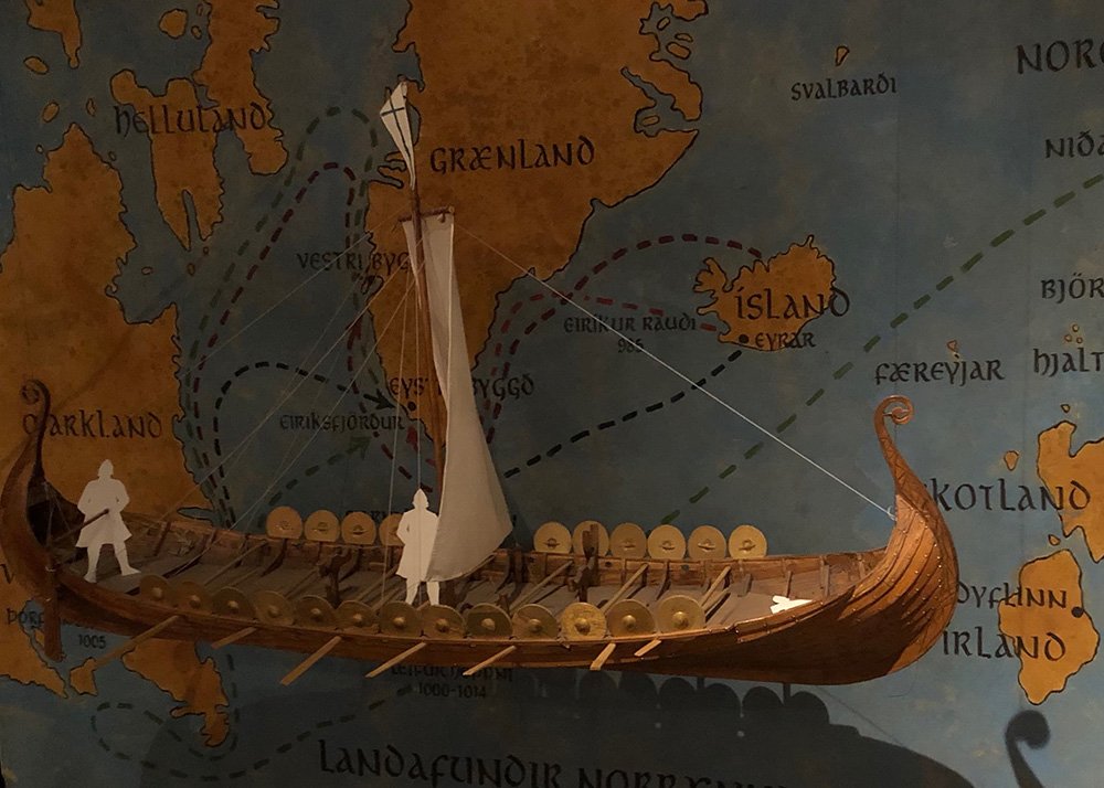 A detailed model of a Viking ship is on display at Reykjavik’s Saga Museum, set against a colorful map showing historical Viking routes across the North Atlantic. The dark indoor setting adds an element of intrigue and historical discovery, perfect for a family museum day.