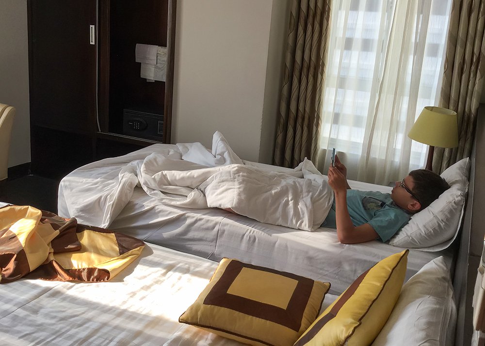 A boy is chilling in a hotel room bed with his ipad. The sun shines through the windows. There's another empty and still made up bed next to him.