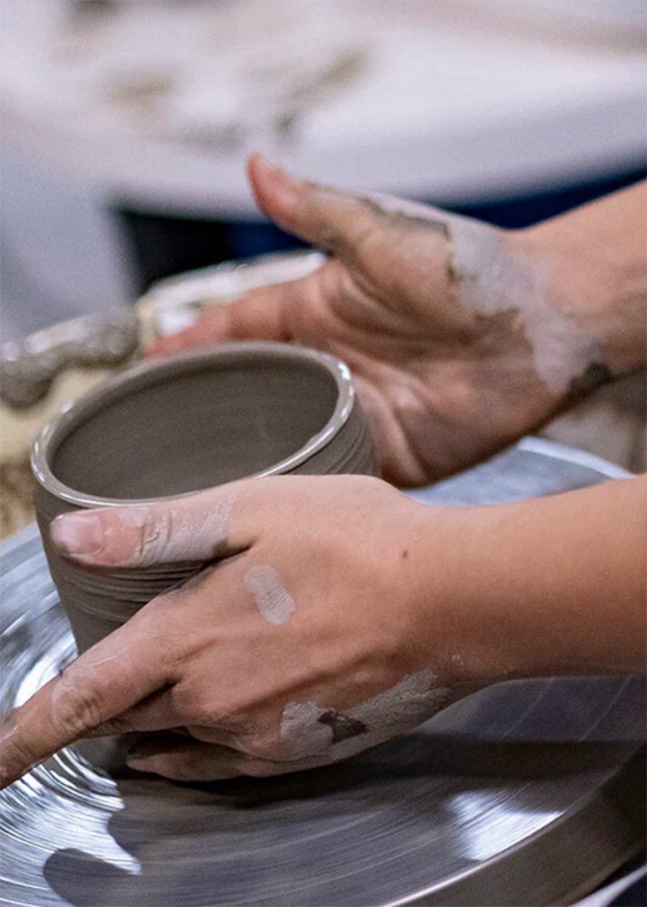 A close-up of a person’s hands shaping clay on a pottery wheel during a pottery class. The hands are coated in clay.