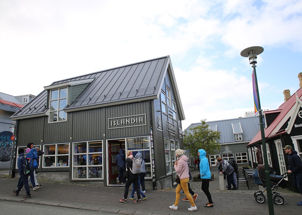 A group of tourists, some with strollers and children, walk down a street in downtown Reykjavik, Iceland. The traditional Icelandic houses with their metal roofs and wooden facades create a charming backdrop. The weather is cool, with people dressed in jackets, reflecting the typical chilly yet refreshing Icelandic climate.