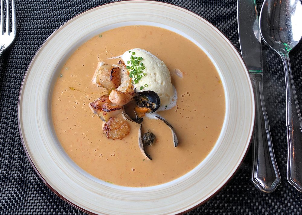 A white plate is filled with a creamy shellfish soup. Mussels, crayfish and a dot of creme fraiche are visible on top.