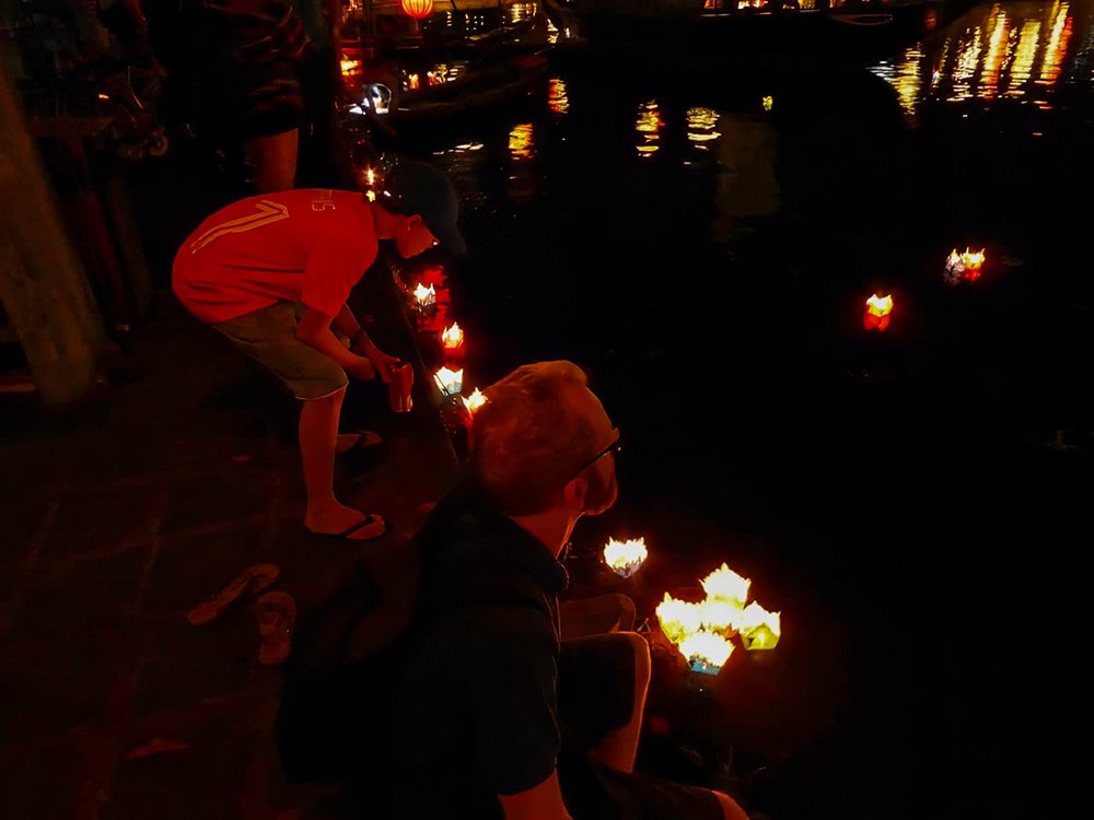 A father and son are standing on the edge of the river in Hoi An at night time after just putting their paper lanterns in the river and making a wish. They're looking at their lanterns in the water.