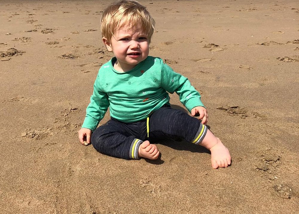 A young toddler in a green shirt and navy pants sits on a sandy beach, looking slightly perplexed by the camera. The child is barefoot, enjoying a quiet moment on a cool, overcast day at an Icelandic beach. Footprints in the sand suggest recent playful activity nearby.