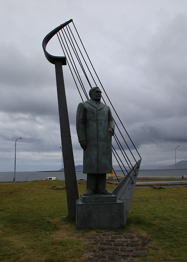 A tall statue of Icelandic poet Einar Benediktsson stands in front of Höfði House in Reykjavik, Iceland, near the seafront. The bronze figure is framed by a harp-like sculpture, set against a cloudy sky and grassy surroundings. The scene evokes both literary and maritime history with distant views of the ocean.