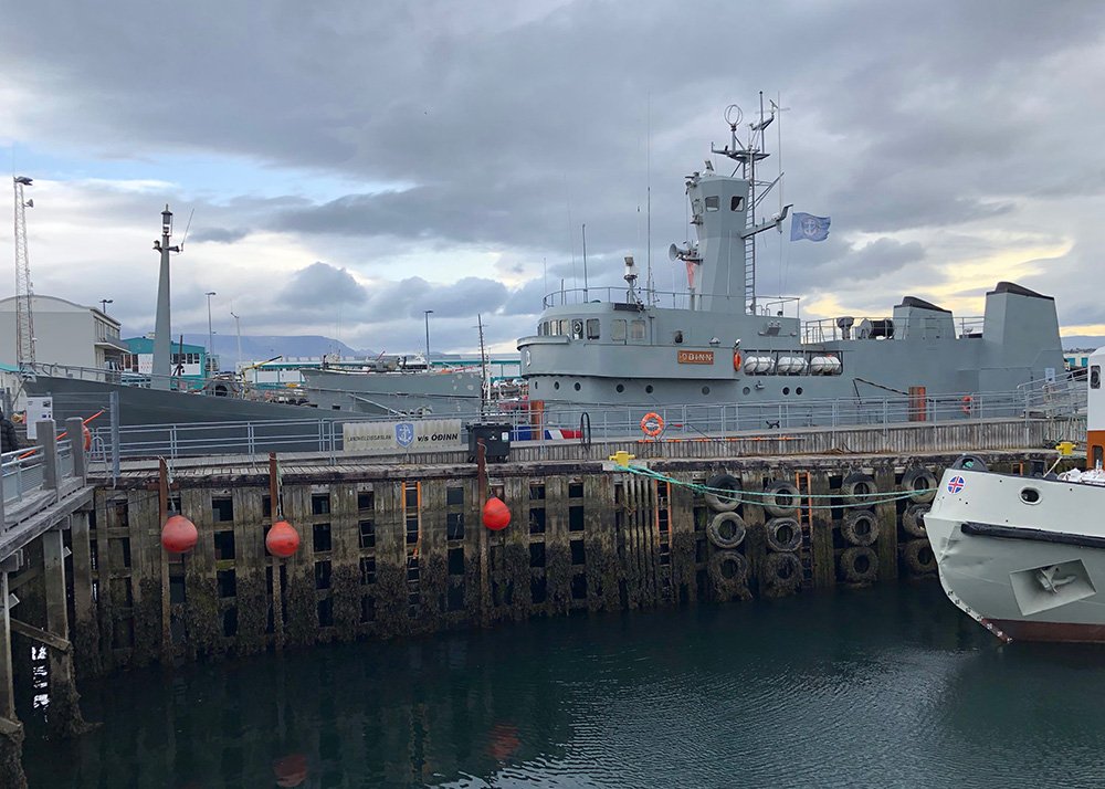 A docked fishing ship, part of the collection at Reykjavik's Maritime Museum, sits in the harbor. The weathered boat, with its white and orange color scheme, evokes a sense of Iceland’s rich maritime history. The overcast sky and calm waters give the scene a quiet, reflective atmosphere.