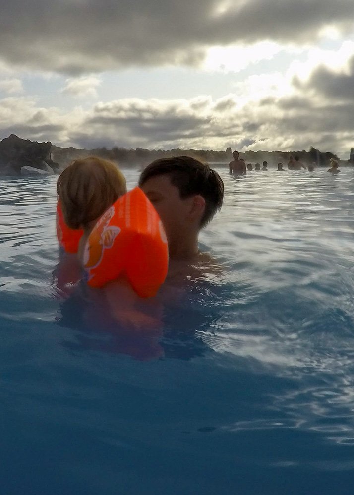 A brother holds his baby brother, who is wearing bright orange floaties, in the warm waters of Laugardalslaug, Reykjavik. The misty horizon and other bathers in the distance give the scene a serene, family-friendly atmosphere, perfect for a relaxing geothermal swim.