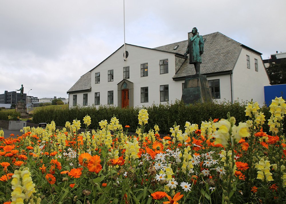 A beautiful white historic building, the Prime Minister's Office, stands in Reykjavik, Iceland, surrounded by a lush garden of vibrant orange and yellow flowers. A statue of Hannes Hafstein, Iceland’s first Prime Minister, stands proudly in front of the building. The sky is cloudy, and the scene exudes peaceful charm in the heart of the city.