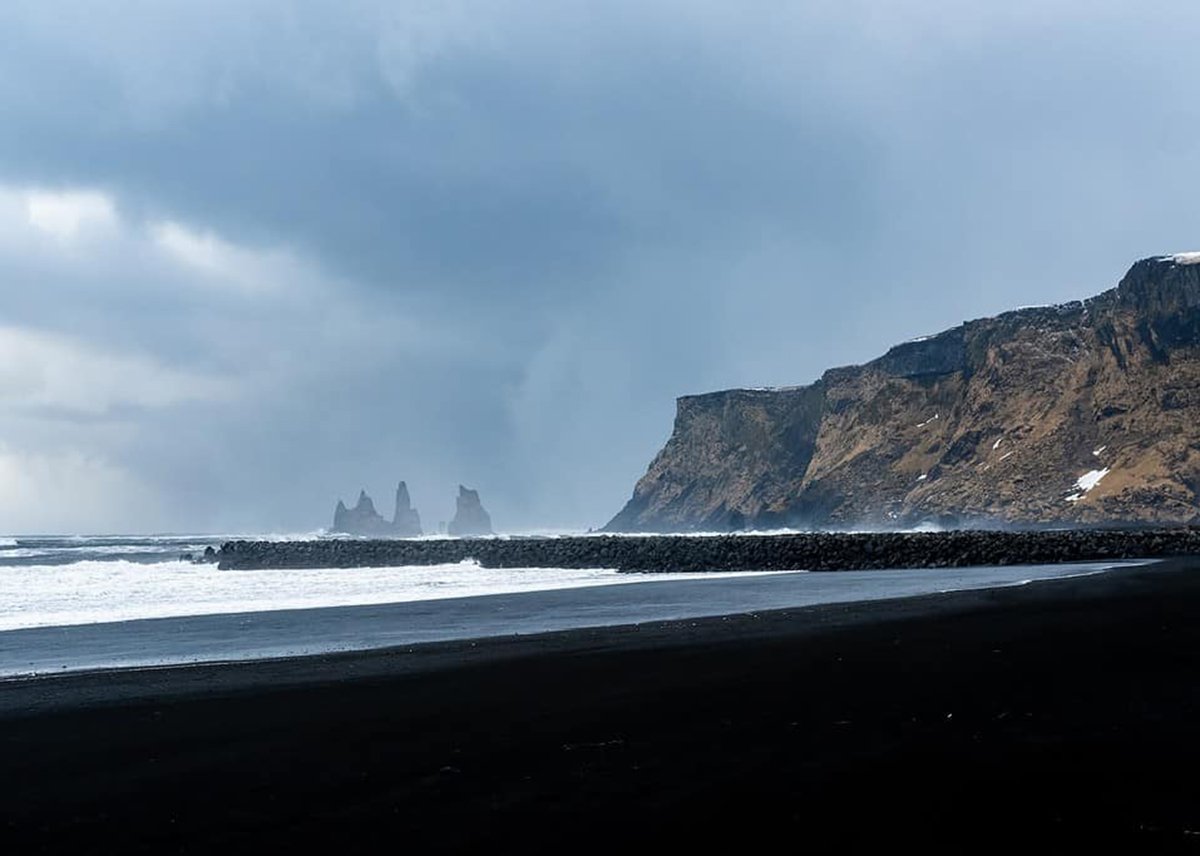 The black sand beach at Reynisfjara with a mountain in the background and the Reynisdrangar basalt sea rock can also be seen in the distance.
