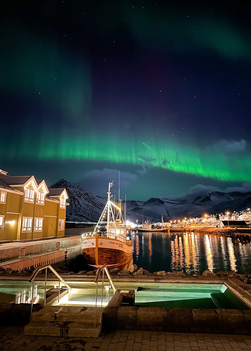 The Siglo Hotel with the harbor in front of it and fisher boats docked. In the background the Northern Lights are dancing and the mountains in the back can be seen, covered in snow.