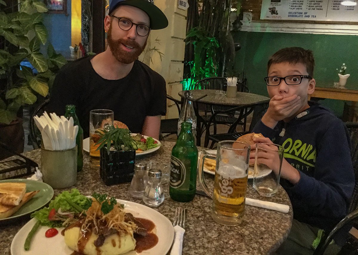 A father and son enjoy a casual dinner at a cozy Hanoi restaurant with burgers, fries, and local beer on the table. A fun and relatable dining moment to highlight when planning a family trip to Hanoi.