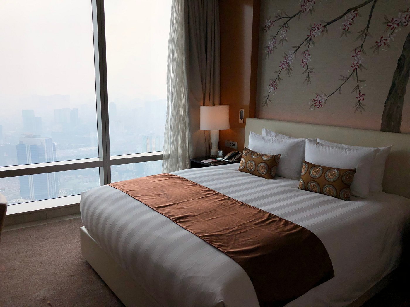 Modern hotel room at Lotte Hotel Hanoi featuring a king-sized bed, large city-view window, and cherry blossom wall art. Ideal lodging inspiration for families planning a family trip to Hanoi.
