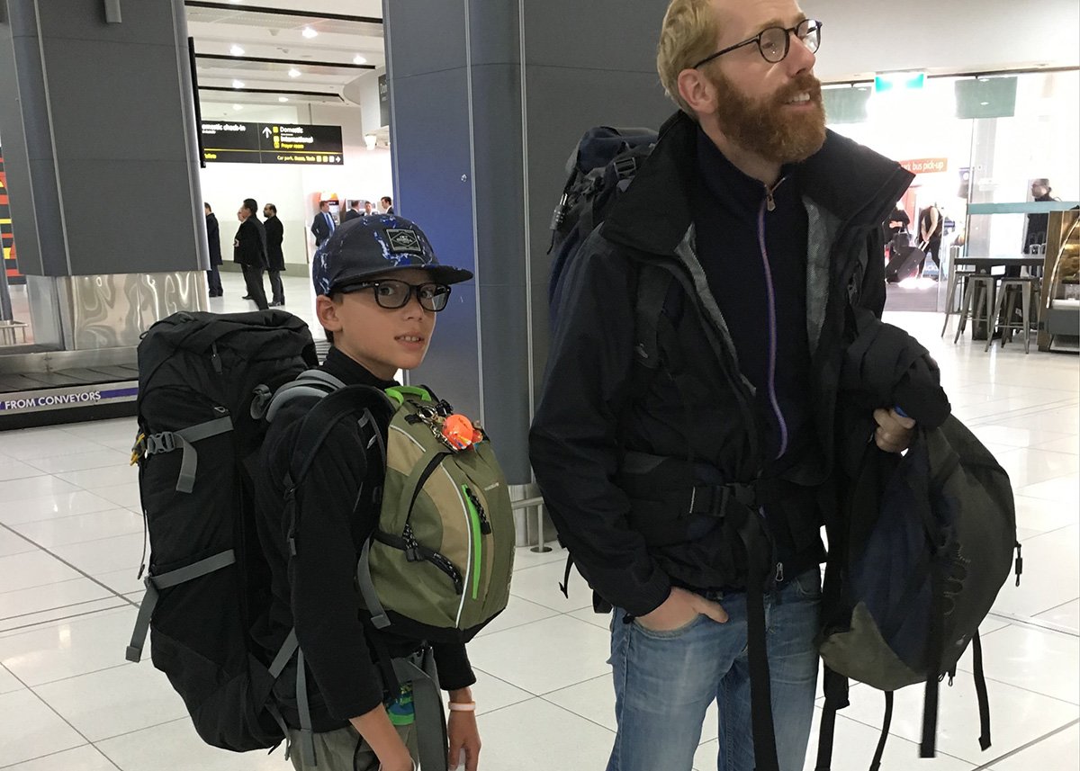 A man and a boy, both wearing large travel backpacks, stand together in a modern airport terminal. The boy, wearing a cap and glasses, looks prepared for an adventure, while the man smiles in a black jacket and jeans, holding an additional bag. Their luggage-heavy look captures the essence of long-term family travel.
