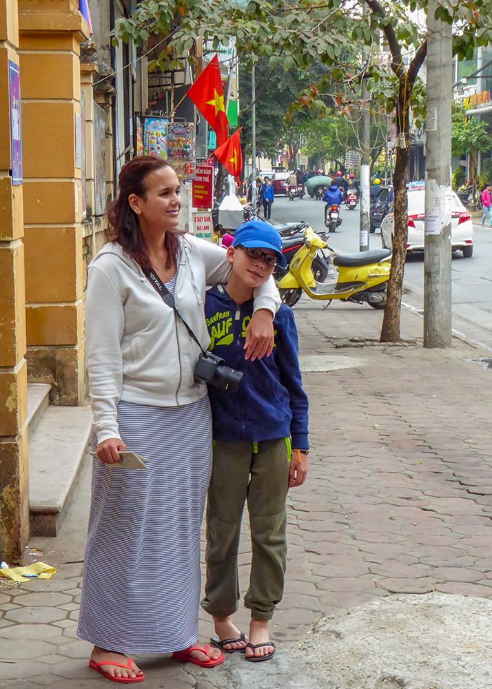 A mother and son stand close together on a bustling Hanoi sidewalk, surrounded by local shops and Vietnamese flags. A heartfelt travel moment that captures the joy of planning a family trip to Hanoi.