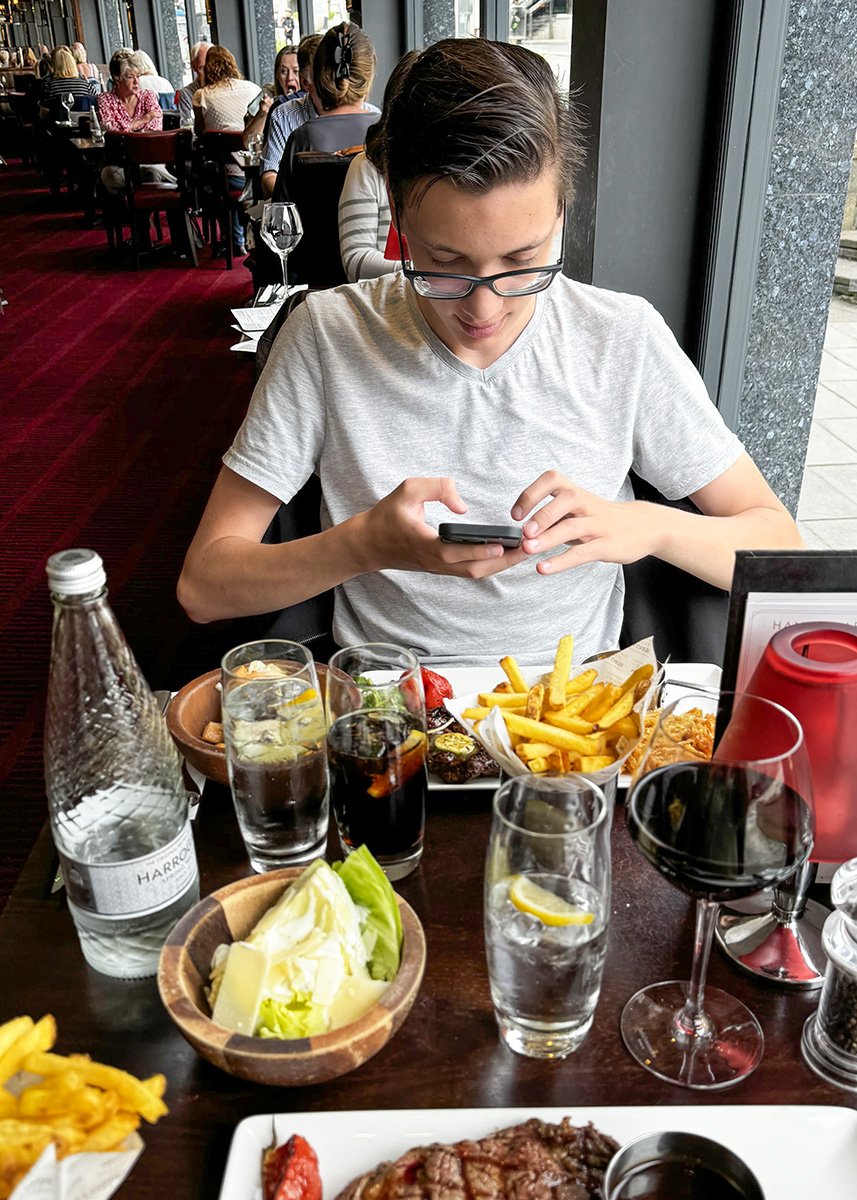A teenage boy in a restaurant taking a picture of his meal: a steak and fries.