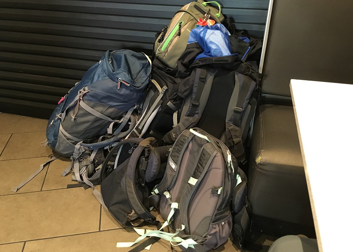 Several large travel backpacks are stacked unattended in a corner booth of a fast-food restaurant. The bags vary in color and size, with one noticeably tagged with green ribbons, suggesting group or family travel. The dim indoor lighting and public location highlight a moment where luggage security could become an issue while traveling.