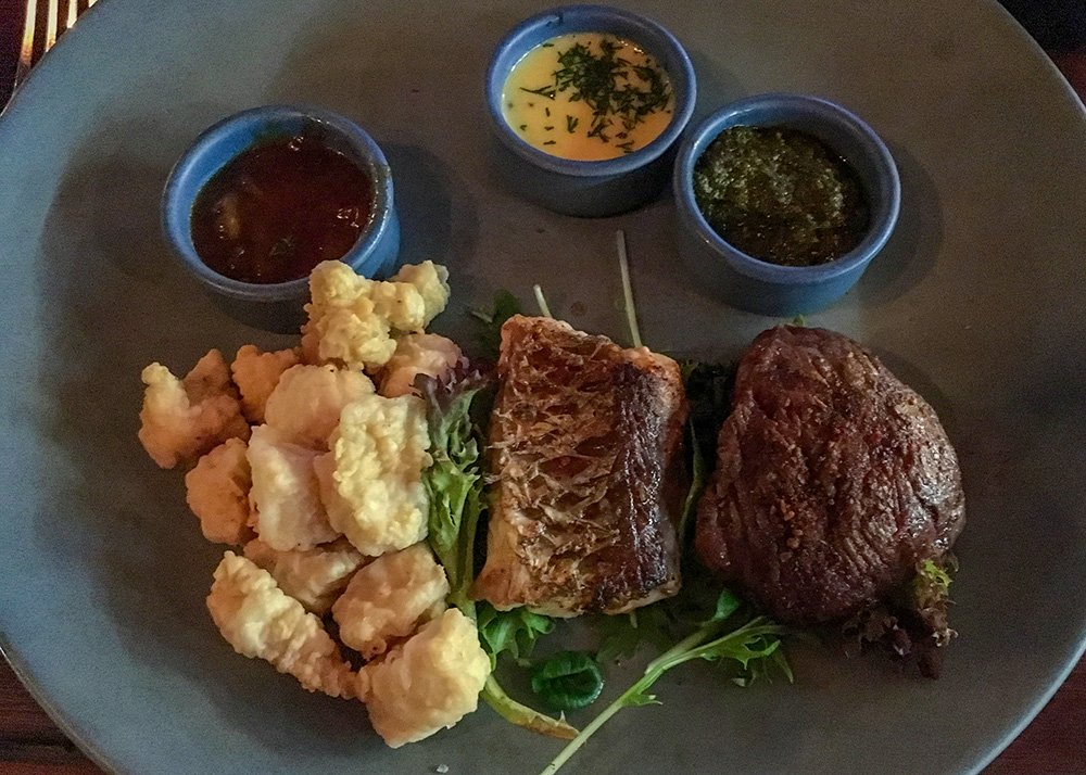 A dark grey plate is filled with a piece of barramundi, kangaroo steak and tater tots. 3 tiny bowls are filled with sauces.