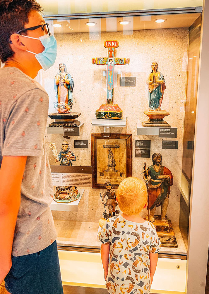 Two children observe colorful Catholic artifacts in a glass case inside a Mérida museum.