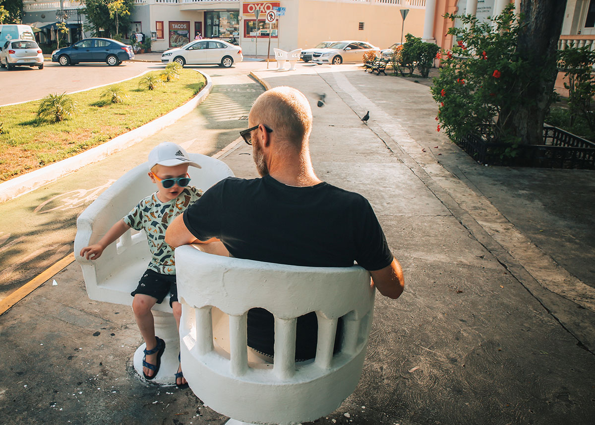 Man and child sit in white “lovers’ chairs” in a sunny plaza in Mérida, near shops and parked cars.