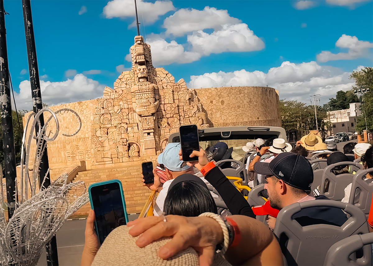Tourists on an open-top bus photograph the intricate stone Monumento a la Patria on Paseo de Montejo in Mérida.