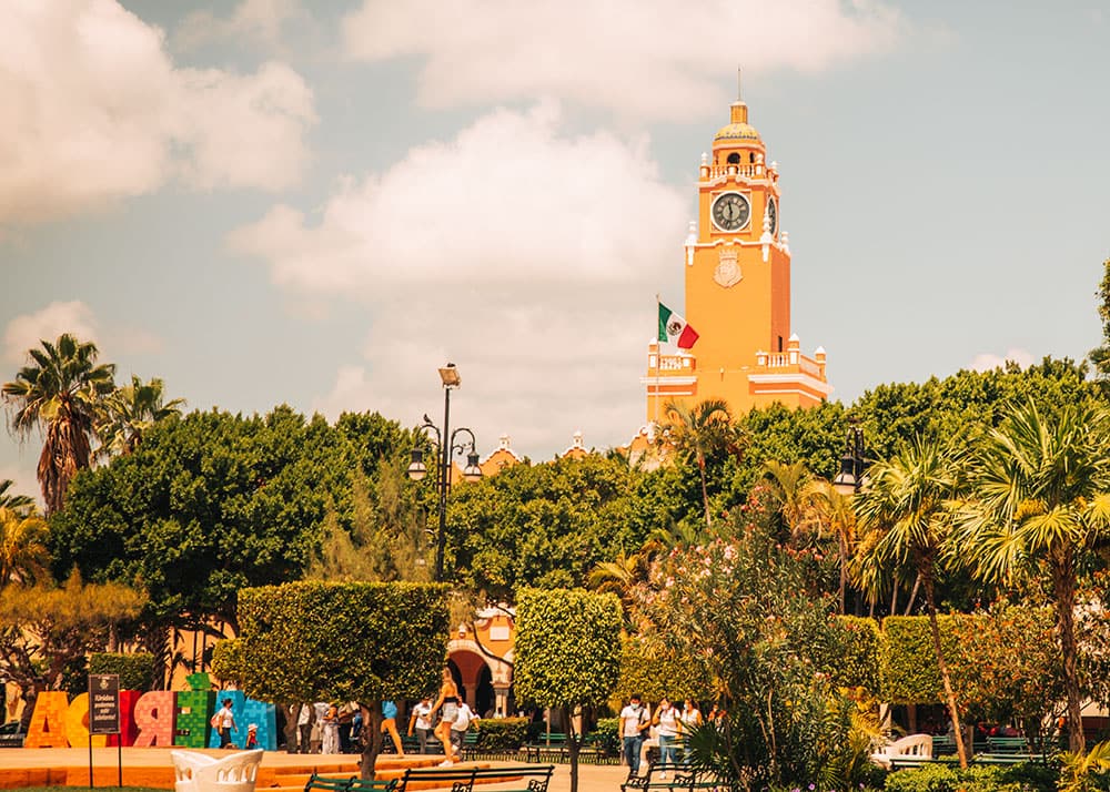 Orange clocktower rises over trees and pathways in Mérida’s Plaza Grande on a sunny day.