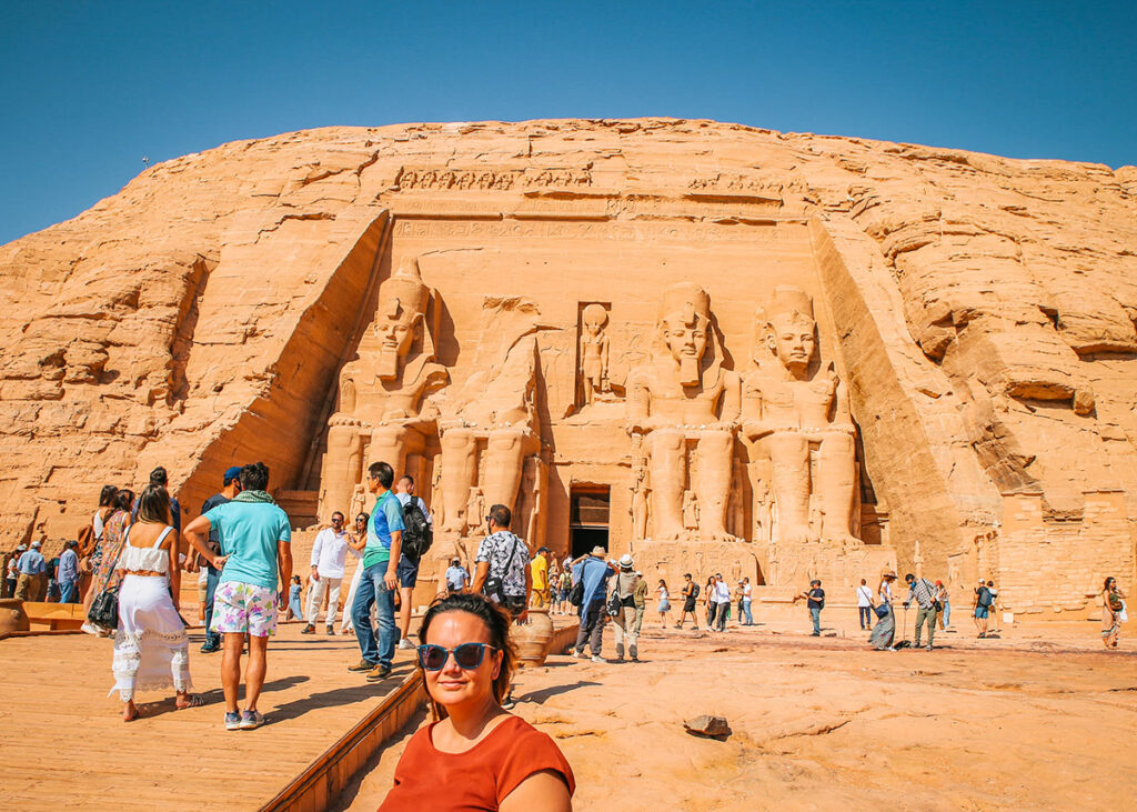 A woman wearing sunglasses and a dark red t-shirt poses in fron of the temple of Ramses II in Abu Simbel