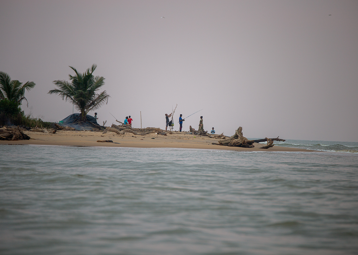 Locals fish with rods on a driftwood-strewn beach in Negombo, beneath a hazy sky.